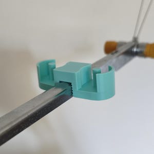 Peut inclure: Un clip en plastique bleu sarcelle clair avec un bloc central carré et deux bras latéraux incurvés fixés sur une barre métallique rectangulaire. Le clip présente un bord intérieur dentelé qui saisit la surface métallique et deux petits ergots.