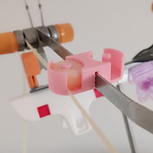 Pode incluir: Close-up de equipamento ortodôntico. Um componente de plástico rosa com um pequeno cilindro de cor clara está preso a um braço de metal. O dispositivo está sobre um fundo branco, com outras partes do equipamento visíveis.