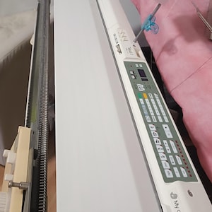 Puede incluir: Máquina de tejer blanca con un panel de control con botones y una pequeña pantalla. La máquina tiene una larga bancada de agujas metálicas y una cubierta blanca. El texto "My creative land" es visible.