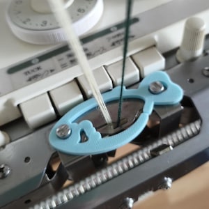 May include: Close-up of a knitting machine with a light blue yarn guide. White and green threads are threaded through the guide. The machine has white keys and a dial with numbers.