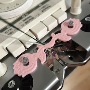 May include: A close-up of a knitting machine with a light pink floral-shaped tool attached. The machine has white buttons and a dial. The tool is secured with silver screws. The background is a light beige.