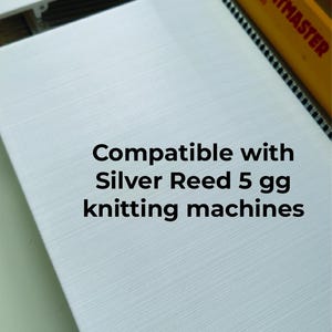 以下が含まれることがあります： テクスチャ加工された白い長方形の編み機用アクセサリー。上部には「Empisal-KNITMASTER MODEL 360」と記された黄色いバーがあります。白い表面には「Compatible with Silver Reed 5 gg knitting machines」というテキストが記載されています。