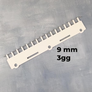 Puede incluir: Un peine de transferencia para máquina de tejer de plástico color crema con una fila de agujas de metal en el borde superior. La herramienta incluye dos orificios circulares y dos ranuras rectangulares, con el texto 9 mm y 3gg impreso en negro.