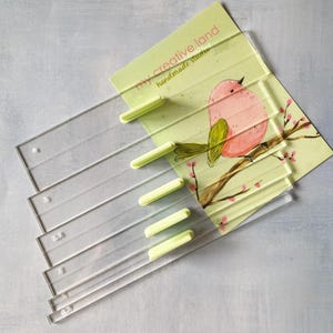 Puede incluir: Reglas de acolchado de acrílico transparente con asas verde claro apiladas junto a una tarjeta con una ilustración de pájaro en acuarela. La tarjeta dice "my creative land handmade studio". Las reglas están marcadas en centímetros.