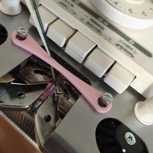 May include: Close-up of a knitting machine with a pink tool, white buttons, and a dial. The machine has Japanese text and a black brush. The dial is white with black numbers.