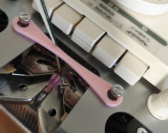 May include: Close-up of a knitting machine with a pink tool, white buttons, and a dial. The machine has Japanese text and a black brush. The dial is white with black numbers.