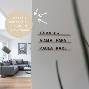 Könnte beinhalten: Eine weiße Wand mit einem Holzschild, auf dem "Familie" mit einem Herzsymbol, "Mama Papa" und "Paula Karl" steht. Das Schild ist aus Holz mit schwarzen Buchstaben. Der Preis gilt pro Buchstabe.