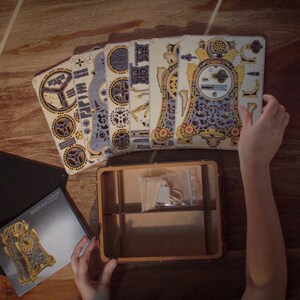 May include: A wooden clock kit with intricate details and a gold and blue color scheme. The kit includes multiple pieces of laser-cut wood and a small box for storing the pieces.