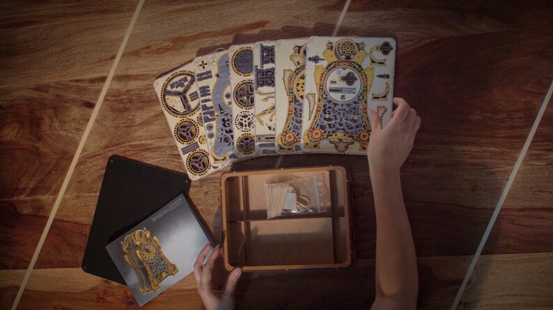 May include: A wooden clock kit with intricate details and a gold and blue color scheme. The kit includes multiple pieces of laser-cut wood and a small box for storing the pieces.