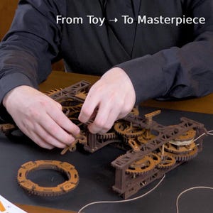 May include: A wooden mechanical clock kit with gears and a circular component. The text "From Toy -> To Masterpiece" is displayed above the clock components. The clock is brown and tan in color.