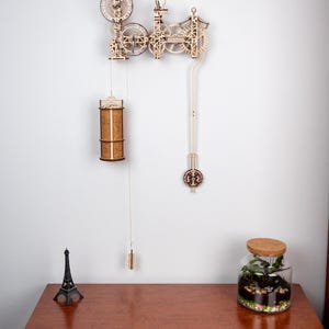 May include: A wooden clock kit with intricate gears and a pendulum. The clock is mounted on a white wall and has a brown wooden finish. The clock is partially assembled and has a small Eiffel Tower figurine on a wooden table below it.