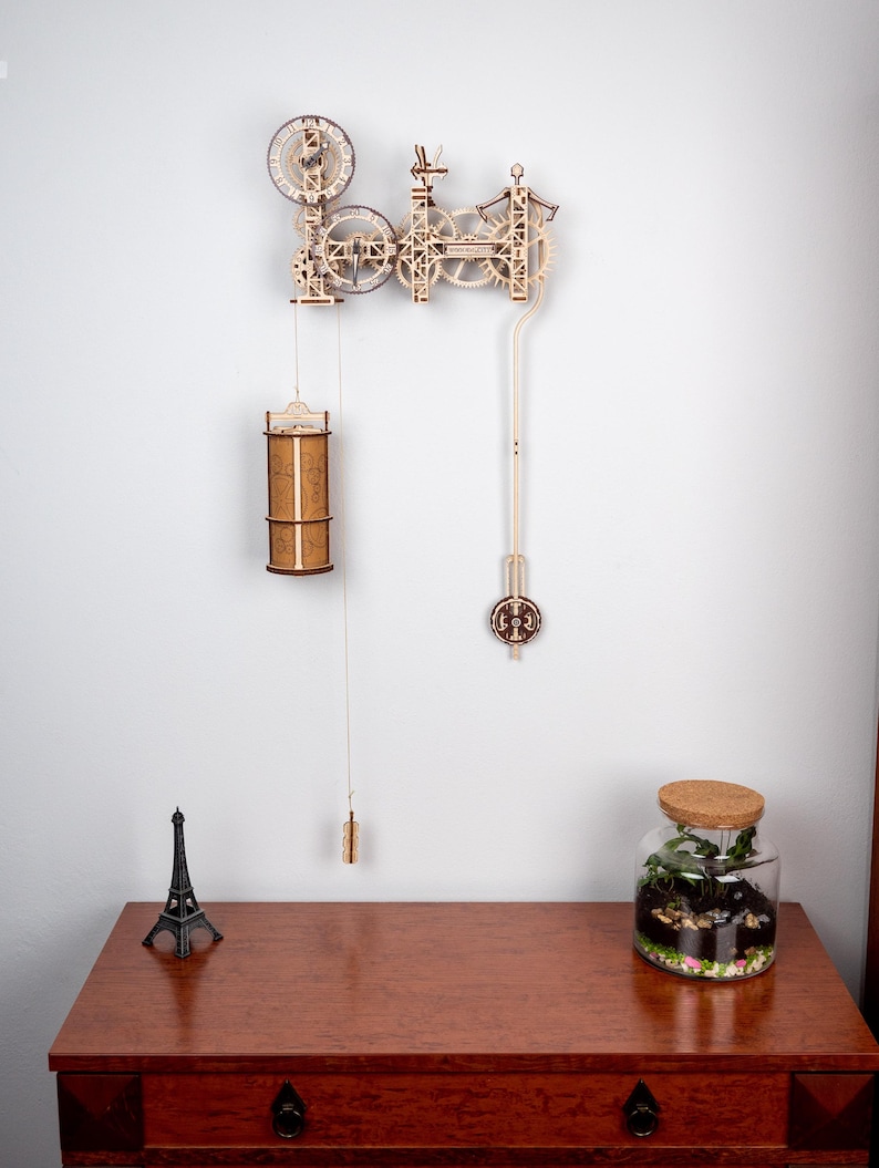 May include: A wooden clock kit with intricate gears and a pendulum. The clock is mounted on a white wall and has a brown wooden finish. The clock is partially assembled and has a small Eiffel Tower figurine on a wooden table below it.