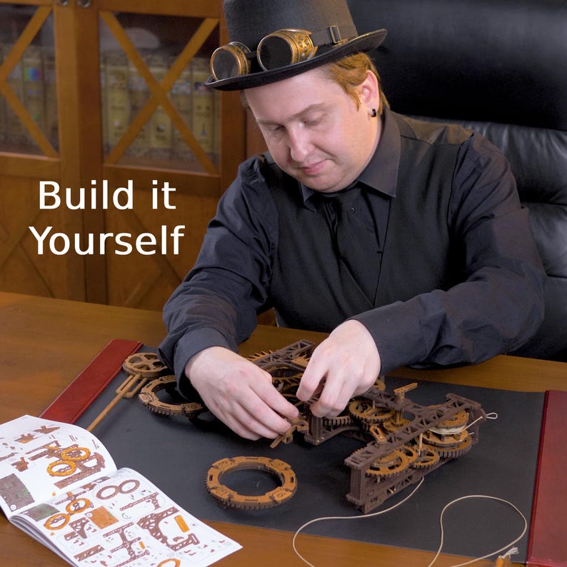 May include: A person assembling a wooden mechanical model with gears and other components. The model is brown and appears to be a clockwork mechanism. The text "Build it Yourself" is visible in the image.