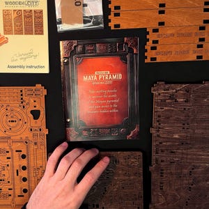 Puede incluir: Un kit de rompecabezas de madera con el texto "MAYA PYRAMID" y "WOODEN CITY" en la portada. El kit incluye piezas de madera, instrucciones de montaje y el texto "Unravel the mystery!". El kit tiene 184 piezas y no requiere pegamento.