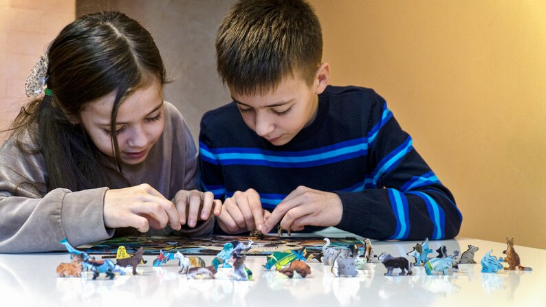 May include: Two children are working on a puzzle with colorful animal figurines. The puzzle is spread out on a white table. The children are focused on the task at hand.