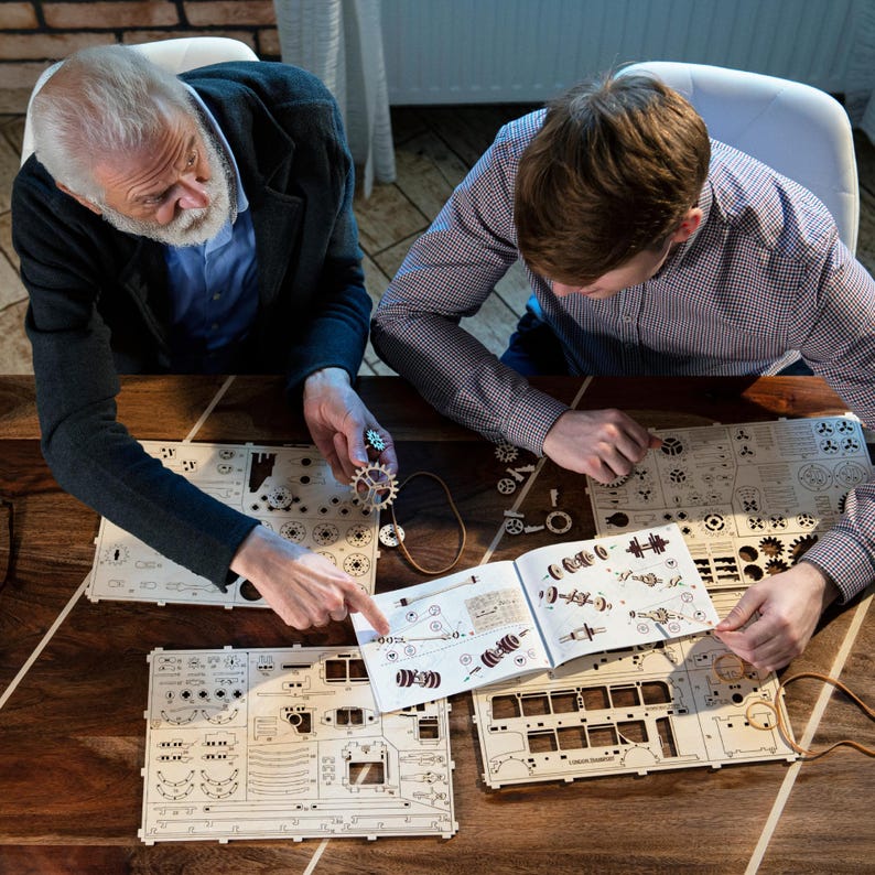 May include: Two people are working on a wooden model kit. The kit includes a variety of gears and other parts. The instructions are laid out on a wooden table.