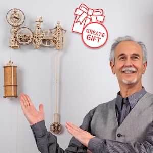 May include: A man smiles and gestures towards a wooden clock kit with intricate gears and a pendulum. The clock is mounted on a white wall. A red and white gift box icon with the text "Great Gift" is in the upper right corner of the image.