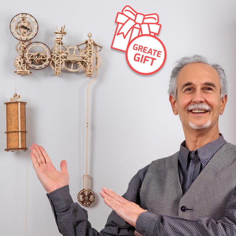 May include: A man smiles and gestures towards a wooden clock kit with intricate gears and a pendulum. The clock is mounted on a white wall. A red and white gift box icon with the text "Great Gift" is in the upper right corner of the image.
