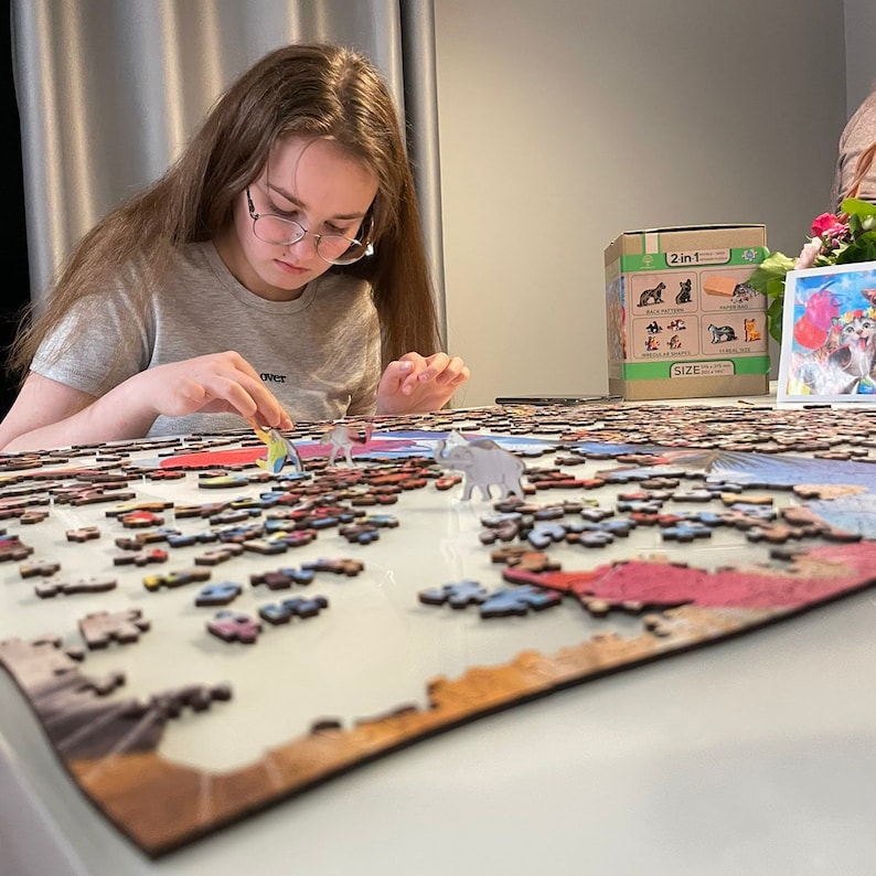 May include: A young person is working on a large jigsaw puzzle with a colorful image. The puzzle pieces are scattered on a table, and the person is carefully placing a piece into the puzzle. There are several small wooden figurines on the table, including a fish, a bird, and an elephant. The puzzle box is visible in the background, and it has a picture of a cat on it. The box says "2 in 1" and "Size: 500 pieces".