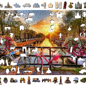 May include: A colorful jigsaw puzzle depicting a canal scene in Amsterdam, Netherlands. The puzzle features a bridge with two bicycles parked on it, a canal with boats, and buildings along the canal. The sky is a vibrant orange and pink, and there are flowers in bloom along the canal.