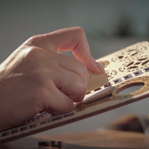 May include: A person's hand is holding a piece of wood with intricate laser cut designs. The wood is light brown with gold accents. The design features a variety of shapes and patterns, including circles, squares, and stars.