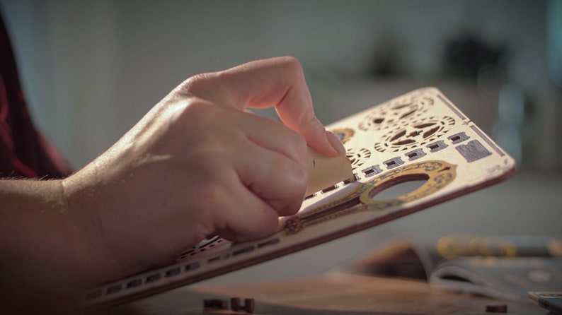 May include: A person's hand is holding a piece of wood with intricate laser cut designs. The wood is light brown with gold accents. The design features a variety of shapes and patterns, including circles, squares, and stars.