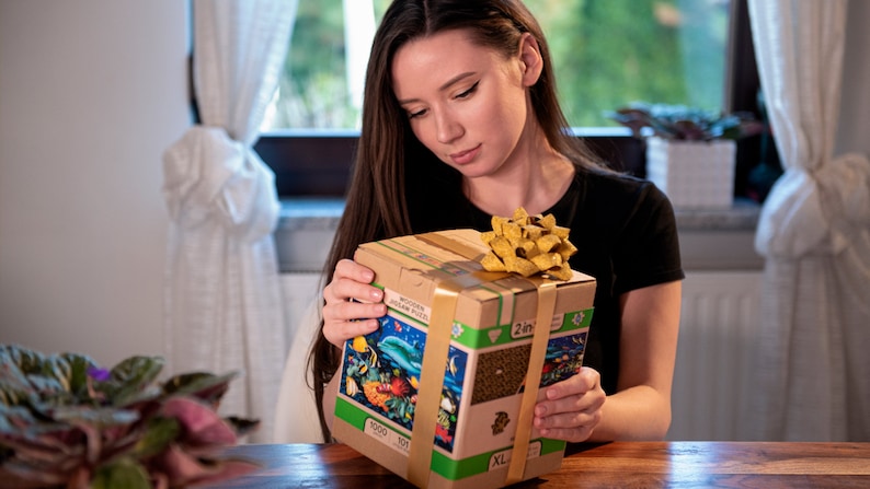 May include: A person is holding a brown cardboard box with a yellow bow. The box has a picture of a colorful underwater scene with fish and coral. The box says "1000+ 2 in 1 XL" on the front.
