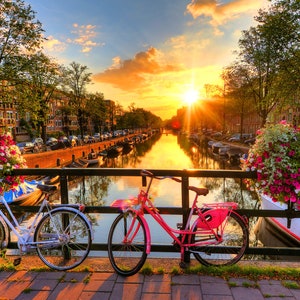 May include: Two bicycles parked on a bridge over a canal in Amsterdam, Netherlands. The bicycles are pink and white, and the canal is lined with buildings and trees. The sun is setting in the background, casting a golden glow over the scene.