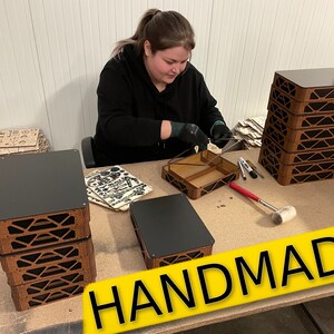 May include: A person assembles wooden boxes with black lids. The boxes are stacked in a pile and labeled "HANDMADE".
