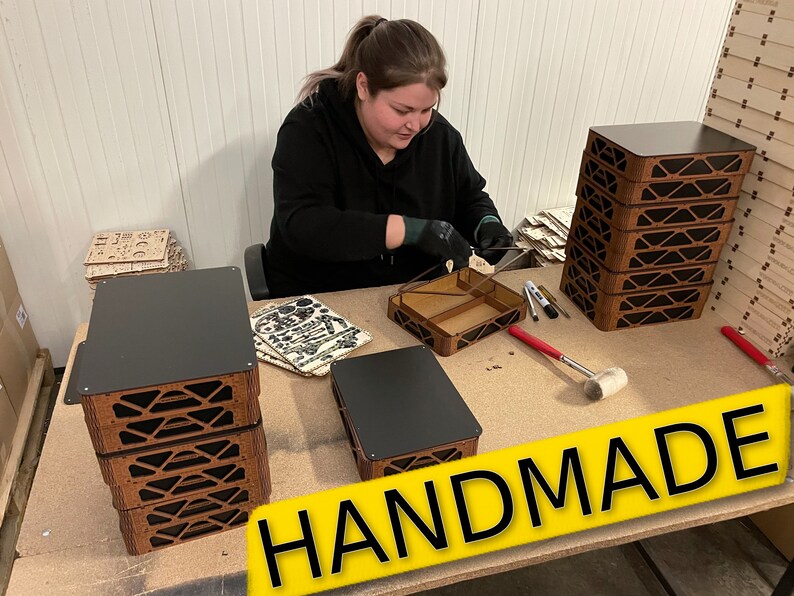 May include: A person assembles wooden boxes with black lids. The boxes are stacked in a pile and labeled "HANDMADE".