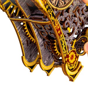 May include: A wooden clock kit with intricate details and gears. The clock face features Roman numerals and a gold-colored frame. The clock is partially assembled and has a hand holding a piece of the clock.
