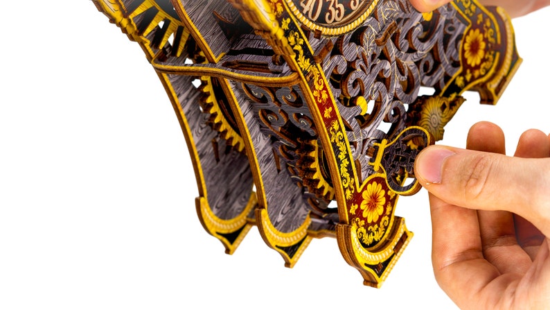 May include: A wooden clock kit with intricate details and gears. The clock face features Roman numerals and a gold-colored frame. The clock is partially assembled and has a hand holding a piece of the clock.