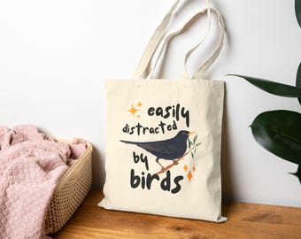 May include: A white tote bag with black text that reads "easily distracted by birds". A black bird with a yellow beak is perched on a branch with green leaves. The bag has a brown handle.