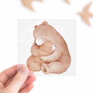 Könnte beinhalten: Aquarellillustration eines Mutterbären, der einen Babybären umarmt, in Brauntönen. Das Bild wird in einer Hand gehalten, mit drei Holzvogel-Ausschnitten im Hintergrund. Das Kunstwerk erinnert an Familie und Geborgenheit.