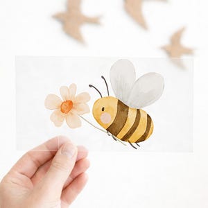Könnte beinhalten: Aquarellillustration einer Biene, die eine Blume trägt. Die Biene ist gelb und braun mit weißen Flügeln. Die Blume ist hellorange. Das Bild wird vor einem weißen Hintergrund mit Holzvogel-Ausschnitten gehalten.