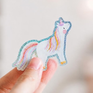 Könnte beinhalten: Ein weißer Einhorn-Aufkleber mit glitzerndem blauem Rand, mit einer mehrfarbigen Mähne und einem Schweif in Rosa, Gelb und Orange, einem kleinen rosa Horn und rosa Wangen, präsentiert als gestanzter Klebeaufkleber mit funkelndem Finish.