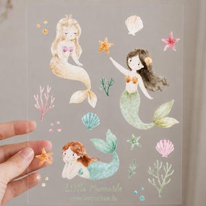 Könnte beinhalten: Ein transparenter Bogen mit Aquarellillustrationen von drei Meerjungfrauen mit blondem, braunem und kastanienbraunem Haar, begleitet von verschiedenen Meeresmotiven wie rosa und blauen Muscheln, orangefarbenen und grünen Seesternen und Korallen.