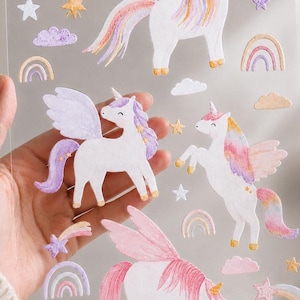 Könnte beinhalten: Ein transparenter Aufkleberbogen mit mehreren Illustrationen von weißen Einhörnern mit bunten Mähnen, Flügeln und Hörnern. Der Bogen enthält zudem verschiedene dekorative Elemente wie pastellfarbene Regenbögen, kleine Wolken sowie gelbe und violette Sterne.