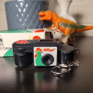 May include: A black and white Kellogg's Corn Flakes Mini 110 camera with a red button and a rooster logo. The camera is attached to a silver keychain.
