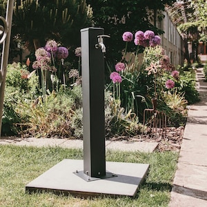 Op de afbeelding: Een zwarte metalen buitenfontein met een kraan, staande op een vierkante betonnen basis in een tuin. De fontein is omgeven door groen gras en paarse bloemen.