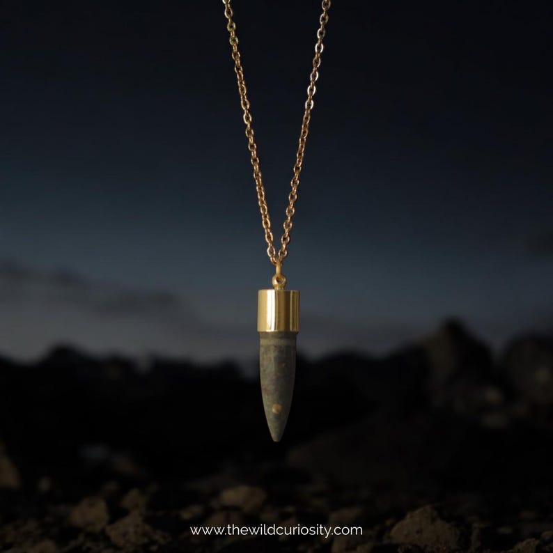 Puede incluir: Un collar de cadena dorada con un colgante con forma de bala. El colgante est&aacute; hecho de metal oscuro con una parte superior dorada.