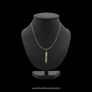 Puede incluir: Un collar de cadena dorada con un colgante de piedra verde en forma de bala. El collar se muestra en un busto de maniqu&iacute; negro.