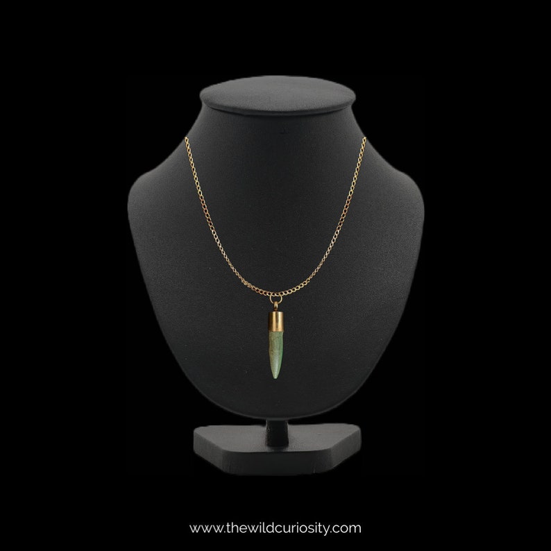 Puede incluir: Un collar de cadena dorada con un colgante de piedra verde en forma de bala. El collar se muestra en un busto de maniqu&iacute; negro.