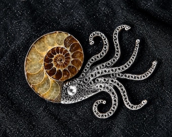 May include: A brooch shaped like an octopus with a spiral ammonite fossil shell head in shades of amber and brown. The body and tentacles are crafted from silver-toned metal with a textured finish and a single clear stone. Text reads thewildcuriosity.com.