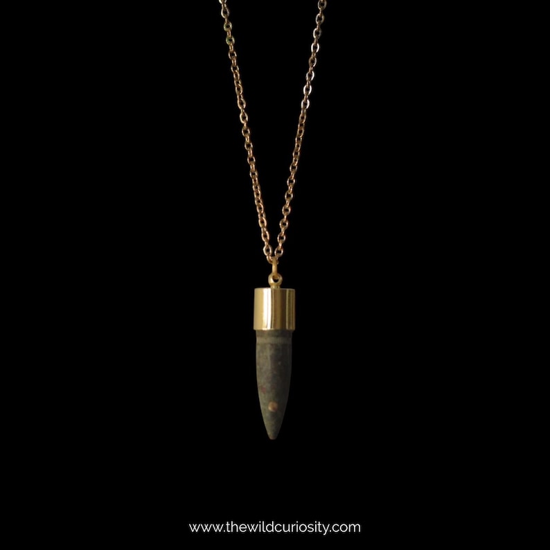 Puede incluir: Un collar de cadena dorada con un colgante de bala de color gris oscuro. El colgante tiene una parte superior e inferior doradas.