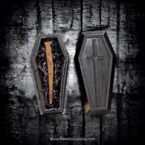 May include: Two miniature black coffin-shaped boxes. One coffin is open, revealing a rusty nail inside, surrounded by dark, dried plant matter. The other coffin is closed, with a cross design on the lid. The website address www.thewildcuriosity.com is at the bottom.
