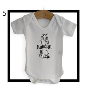 May include: White baby bodysuit with short sleeves, hanging on a wooden hanger. The bodysuit features the text "Cutest Pumpkin in the Patch" in black script, with a pumpkin graphic above the text. The bodysuit is designed for infants.