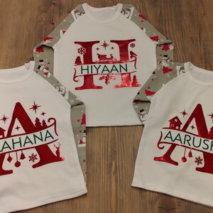 Op de afbeelding: Drie witte shirts met lange mouwen met grijs-witte gepatroneerde mouwen. Elk shirt heeft een rode glitterletter met een kerstthema en een naam: Aahana, Hiyaan en Aarushi.