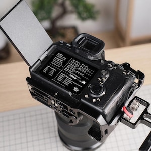 May include: Black digital camera with a lens attached, featuring a flip-out screen displaying settings. The camera has a black cage and a red accent. The screen displays text including "SHUTTER" and "WHITE BALANCE".