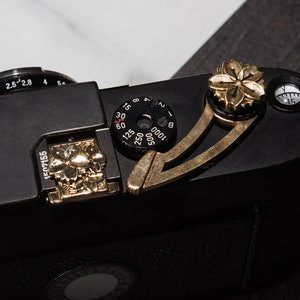 May include: Black camera with gold-colored accents, including a flower-shaped button and a dial. The camera has a leather strap attached. The top of the camera features a dial with numbers and settings. The camera is on a light and dark surface.